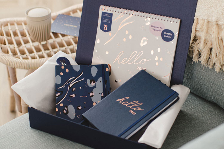 An open gift box on top of a grey sofa.  Inside the box is a navy and pink animal print notebook, a navy diary with "hello 2022" on the middle in rose gold foil and a 2022 calendar with the same foil writing. Part of our new stationery launch