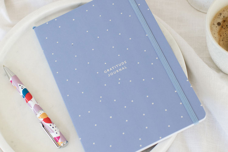 Image of lilac gratitude journal laying on a clothed surface with a brightly coloured pen to the bottom left hand side.  The journal cover has white small dots and in the centre says "gratitude journal" in all caps. Part of our new stationery release
